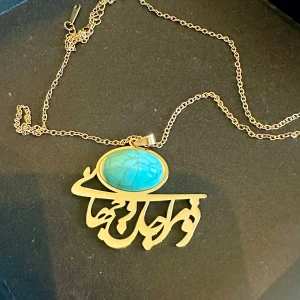 Guldfärgat halsband med turkos sten och persisk text - Unikt halsband med en stor oval turkos sten infattad i guldfärgad metall och ett hänge med dekorativ persisk text.  ”Du är mitt liv och min värld” – dessa ord kommer från den persiske poeten Rumi, och bär på en tidlös kärlek. Ett halsband för dig som vill bära en bit poesi nära hjärtat.