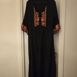 Svart långklänning med broderade detaljer - Svart långklänning med vida ärmar och v-ringning. Klänningen har vackra broderade blommor i orange och blått längs ärmar och framtill. Perfekt för dig som gillar bohemisk stil och vill sticka ut med snygga detaljer.