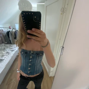 Jeans korsettopp med knappar - Säljer en snygg axelbandslös korsettopp i jeansmaterial med knappar framtill. Toppen har en tight passform och markerad midja, perfekt för att skapa en cool och trendig look.