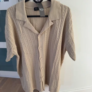 Beige Stickad kofta  - Säljer en beige stickad kofta från Boohoo med korta ärmar och knappar framtill. Tröjan har ett klassiskt kabelstickat mönster och en mjuk krage. Perfekt för lager-på-lager och stilrena outfits. Den är storlek S men är oversized, alltså passar bättre för er som har M/L