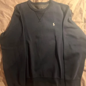 Mörkblå sweatshirt från Ralph Lauren - Mörkblå sweatshirt från Ralph Lauren med klassisk rund halsringning och broderad logga på bröstet. Tröjan har en enkel och stilren design, perfekt för en avslappnad look.