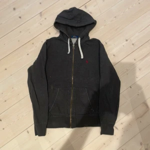 Mörkgrå hoodie från Polo Ralph Lauren - Säljer en mörkgrå hoodie från Polo Ralph Lauren med dragkedja och vit dragsko. Tröjan har huva, två fickor framtill och den klassiska röda broderade loggan på bröstet. Perfekt för en avslappnad stil. Skick 7/10, det är bara att skicka om ni undrar något!