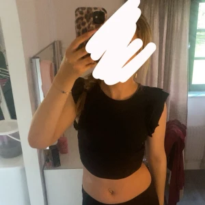 Svart volang topp - Snygg svart croppad topp med volangdetaljer på ärmarna. Perfekt till både jeans och kjol för en trendig look. Mjuk och bekväm passform med rund halsringning.