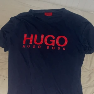t-shirt från Hugo Boss med röd logga - Säljer en mörkblå t-shirt från Hugo Boss med stor röd HUGO-logga på bröstet. Klassisk passform och rund halsringning. Perfekt för dig som gillar stilrena märkesplagg.