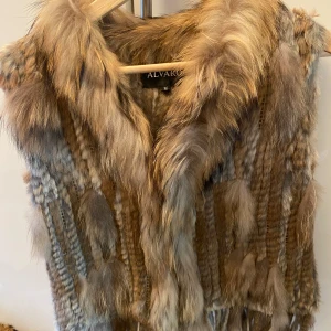 Brun pälsväst från Alvako - Säljer en lyxig brun pälsväst från Alvako i äkta kaninpäls med tvättbjörnspäls vid kragen. Västen har en fluffig och mjuk känsla, perfekt för att addera lite extra stil till din outfit. Fodret är i skinn och pälsen har naturliga färgskiftningar.
