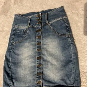 Jeanskjol med knappar framtill - Säljer en klassisk blå jeanskjol med knappar hela vägen framtill och två fickor. Kjolen har en hög midja och är i en kort modell. Perfekt att matcha med en enkel topp eller hoodie.