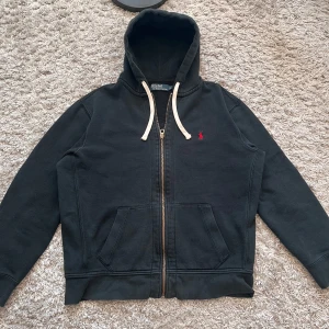 Svart hoodie från Polo Ralph Lauren - Svart hoodie från Polo Ralph Lauren med dragkedja och huva. Tröjan har vita snören, två fickor fram och en liten röd broderad logga på bröstet. Perfekt för en avslappnad stil. Storlek på etiketten är XL men passar bättre L eller M. Se mått.          Armhåla-armhåla: ca 60cm. Längd från axel: ca 62cm.