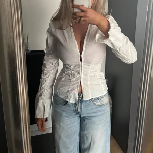 Vit v-ringad blus med dragkedja - Säljer en vit v-ringad blus med dragkedja framtill och markerad midja. Blusen har långa ärmar med breda manschetter och en figurnära passform. Storlek XS 🌸