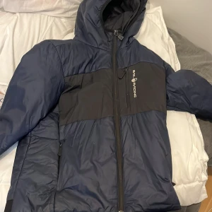 Blå och svart jacka från Sail Racing - Säljer en blå och svart jacka från Sail Racing med huva och dragkedja framtill. Jackan har sportig design och logga på bröstet. Perfekt för kyligare dagar och har en modern look.