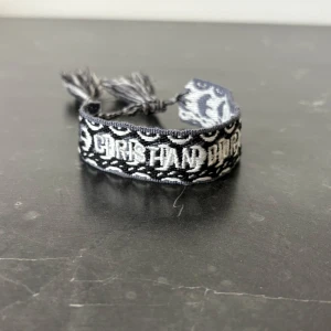 broderat armband från Dior - Säljer ett snyggt svart och vitt armband från Dior med broderad text och mönster. Armbandet har justerbara band med tofsar i ändarna och är perfekt som accessoar för att lyfta din stil.