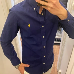 Mörkblå Skjorta från Polo Ralph Lauren - Säljer en klassisk mörkblå skjorta från Ralph Lauren med vit knappstängning och gul broderad logga på bröstet. Skjortan har button-down krage och långa ärmar. Perfekt till dig som gillar stilrena plagg med snygga detaljer. Det står att det är storlek small fast skjortan passar mer som Medium eller Large. Snabb Leverans!
