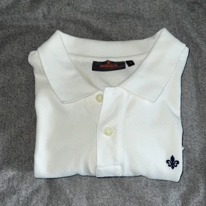 Perfekt Vit pikétröja från Morris - Vit pikétröja från Morris med klassisk krage och två knappar framtill. Liten broderad logga i form av en mörkblå lilja på bröstet. Perfekt basic-plagg med stilren design. Fungerar perfekt nu till sommaren och är praktiskt taget oanvänd typ. Storlek M och true to size därav för stor :(