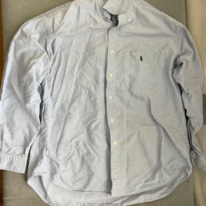 Ljusblå skjorta från Ralph Lauren - Klassisk ljusblå skjorta från Ralph Lauren med broderad logga på bröstfickan. Skjortan har knappar framtill och är tillverkad i 100% bomull. Perfekt för en clean och stilren look.
