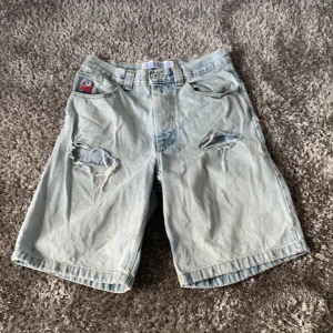 Ljusblå jeansshorts från Big Boy - Säljer ett par ljusblå jeansshorts från Big Boy med slitna detaljer och hål på framsidan. Shortsen har en broderad figur vid fickan och klassisk femficksdesign. Perfekta för en avslappnad stil.
