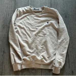 Beige sweatshirt från Polo Ralph Lauren - Klassisk beige sweatshirt från Polo Ralph Lauren med rund halsringning och broderad logga på bröstet. Tröjan har långa ärmar och ribbade muddar vid ärmslut och nederkant. Perfekt för en avslappnad stil.