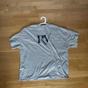 Grå t-shirt från Reclaimed Vintage - Säljer en grå t-shirt från Reclaimed Vintage med stor RV-logga och text på ryggen. T-shirten har en klassisk passform och är tillverkad i mjuk bomull. Perfekt för en avslappnad stil.