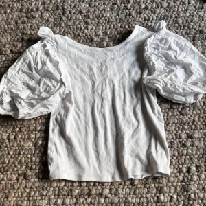 Vit topp med puffärmar från Zara - Säljer en vit ribbad topp från Zara med stora puffärmar. Toppen har rund halsringning och är tillverkad i ett mjukt material. Perfekt för dig som gillar trendiga detaljer och en feminin stil.