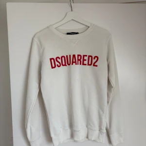 Vit sweatshirt från Dsquared2 - Säljer en vit sweatshirt från Dsquared2 med röd logotyp på bröstet. Tröjan har rund halsringning, långa ärmar och ribbade muddar vid ärmslut och nederkant. Perfekt för en stilren och avslappnad look.