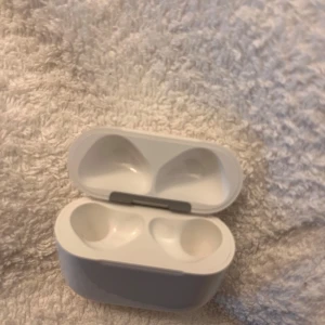 Vit laddningsetui till trådlösa hörlurar - Säljer ett vitt laddningsetui till trådlösa hörlurar. Etuiet har en smidig magnetisk öppning, LED-indikator på framsidan och laddningsport på undersidan. Perfekt för att hålla dina hörlurar laddade och skyddade.