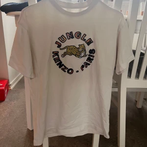 Sällsynt Kenzo jungle t-shirt - Vit t-shirt från Kenzo med färgglatt tryck av en tiger och texten 'Jungle Kenzo Paris' på bröstet. Klassisk passform och rund halsringning. Perfekt för dig som gillar statement-plagg och streetstyle.