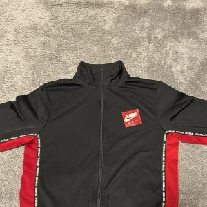 Svart och röd Jordan set - Säljer en svart och röd Jordan Jumpman jacka med dragkedja och hög krage. Jackan har röda paneler på sidorna och vita logotexter längs ärmarna. Jumpman-logga på bröstet. Perfekt för sport eller streetwear. Det är en liten skada på ena byxbenet men annars är det bra skick!
