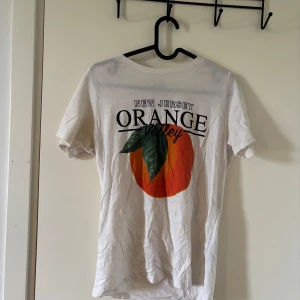 Vit t-shirt med New Jersey Orange-tryck - Säljer en vit t-shirt med stort tryck på ryggen: 'New Jersey Orange Valley' och en stor apelsin med gröna blad. T-shirten har rund hals och korta ärmar. Perfekt för dig som gillar fruktiga prints och vill sticka ut lite extra.