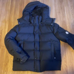 Svart dunjacka från Moncler med huva - Säljer en svart dunjacka från Moncler med huva och stora, quiltade partier. Jackan har långa ärmar, knappar och dragkedja framtill samt fickor på sidorna. Klassisk Moncler-logga på ärmen och innerfoder med serietidningsmotiv.