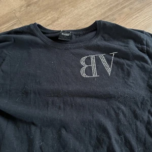 Svart t-shirt med strasstext från Blacki - Svart t-shirt med glittrande strasstenar som bildar texten 'VB' fram och 'NEED MONEY FOR PORSCHE' bak. Rund halsringning och korta ärmar. Perfekt för dig som gillar statement-plagg med lite attityd.