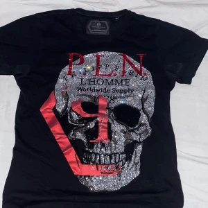 Philipp plein  - Cool svart t-shirt från Plein Sport med stort glittrigt dödskalletryck och röda detaljer framtill. T-shirten har rund hals och korta ärmar, perfekt för en edgy streetstil. Äkta såklart:)