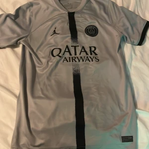PSG Mbappé fotbollströja grå - Säljer en grå PSG fotbollströja med svart vertikal rand, Paris Saint-Germain-logga och Jordan-märke på bröstet. Qatar Airways som sponsor framtill och Mbappé 7 på ryggen. Perfekt för fans av både PSG och Mbappé.