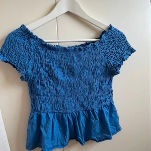 Blå smockad topp med volang - Säljer en blå smockad topp med korta ärmar och volang nertill. Toppen har en elastisk, rynkad överdel och en söt, avslappnad passform. Perfekt för varma dagar!