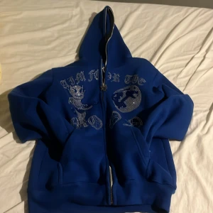 Blå hoodie med strasstenar och tryck - Säljer en blå hoodie med dragkedja och huva. Framsidan har ett stort tryck med strasstenar, inklusive en tecknad figur och texten 'AIM FOR THE MOON'. Perfekt för dig som gillar streetwear och vill sticka ut.