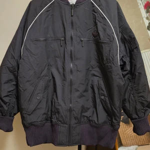 Svart bomberjacka från Adidas - Säljer en svart bomberjacka från Adidas med vita detaljer längs axlarna och ribbade muddar. Jackan har dragkedja framtill, två bröstfickor med dragkedja och två snedställda fickor med knapp. Klassisk Adidas-logga på bröstet. Passar till både Man och Kvinna.