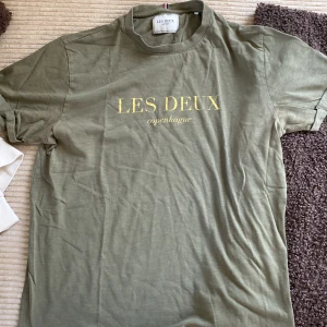 Olivgrön t-shirt från Les Deux - Snygg olivgrön t-shirt från Les Deux med gul text på bröstet. Klassisk passform och rund halsringning. Perfekt för en avslappnad stil och enkel att matcha med jeans eller shorts.