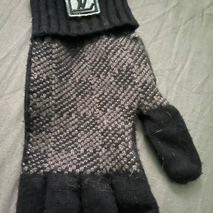 Svarta stickade handskar från Louis Vuitton - Stickade svarta handskar med gråmelerat mönster och ribbade muddar. Handskarna har en tydlig LV-logga på mudden och är perfekta för kyliga dagar.