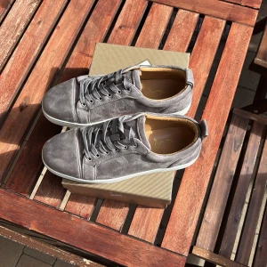 Gråa sneakers från Christian Louboutin - Säljer ett par gråa sneakers från Christian Louboutin med klassisk röd sula och snörning. Skorna är i mocka med vita gummisulor och har rund tå. Perfekta för dig som vill ha något stilrent men ändå sticker ut.