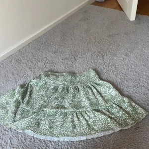 Grön blommig kjol från Gina Tricot - Söt grön kjol från Gina Tricot med vitt blommönster och volangkant. Kjolen har resår i midjan och är lätt och luftig, perfekt för varmare dagar.