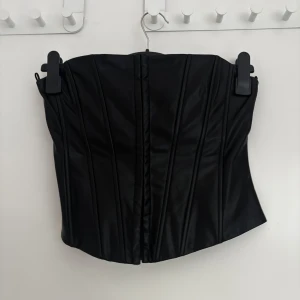 Svart korsett-topp i skinnimitation - Säljer en svart korsett-topp i skinnimitation med markerade sömmar för en snygg siluett. Toppen är axelbandslös och har en stilren, modern look. Perfekt för att lyfta vilken outfit som helst.