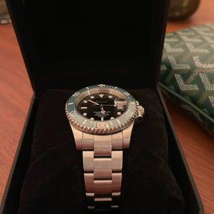 Silverfärgad Seiko herrklocka med grön bezel - Stilren Seiko herrklocka i silver med grön bezel och svart urtavla. Klockan har datumvisning och tydliga indexmarkeringar. Länkarmband i rostfritt stål och klassisk rund form. Perfekt till sommaren. Storlek: 40mm Utseende: Mörk urtavla med grön ring Glas: Safirglas med inzoomad datumvisare Baksida: Öppen baksida (genomskinlig bakboett) Visare: Självlysande visare och ur som lyser i mörker Armband: Oyster eller jubilee-armband i borstat stål  Urverk: Automatiskt Seiko NH35 med 41 timmars. 