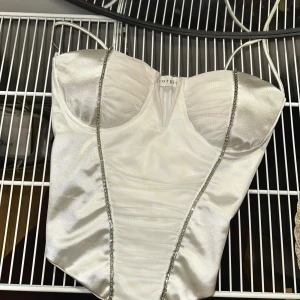 Vit satin korsettopp med glittriga axelband från Giorgia - Vit korsettopp i satin med glittriga axelband. Toppen har vadderade kupor och smickrande sömmar framtill. Köpt ifrån raglady, använd 2 gånger. Eftersom tyget är satin så är den väldigt känslig.