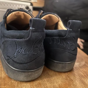 Mörkblå sneakers från Christian Louboutin - Säljer ett par mörkblå sneakers från Christian Louboutin i mocka med klassisk röd sula och diskret broderad logga på hälen. Skorna har rund tå och svarta snören. Insidan är ljusbeige med guldfärgad logga.