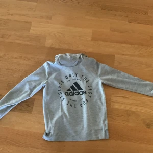 Grå hoodie från Adidas - Säljer en stilren grå hoodie från Adidas med tryck på framsidan. Tröjan har långa ärmar och en bekväm huva. Perfekt för en avslappnad stil.