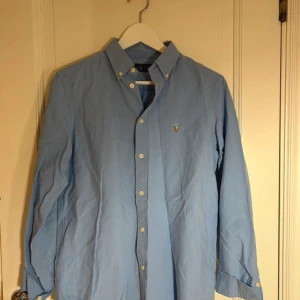 Blå skjorta från Ralph Lauren - Snygg blå skjorta från Ralph Lauren med klassisk krage och broderad logotyp på bröstet. Skjortan har långa ärmar och knappar. 