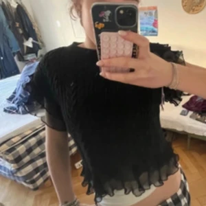 Svart croppad topp med volang - Säljer en svart croppad topp med volangkant nertill och vid ärmslut. Toppen har korta ärmar och är tillverkad i ett lätt, syntetiskt material. Perfekt för dig som gillar trendiga och feminina detaljer.