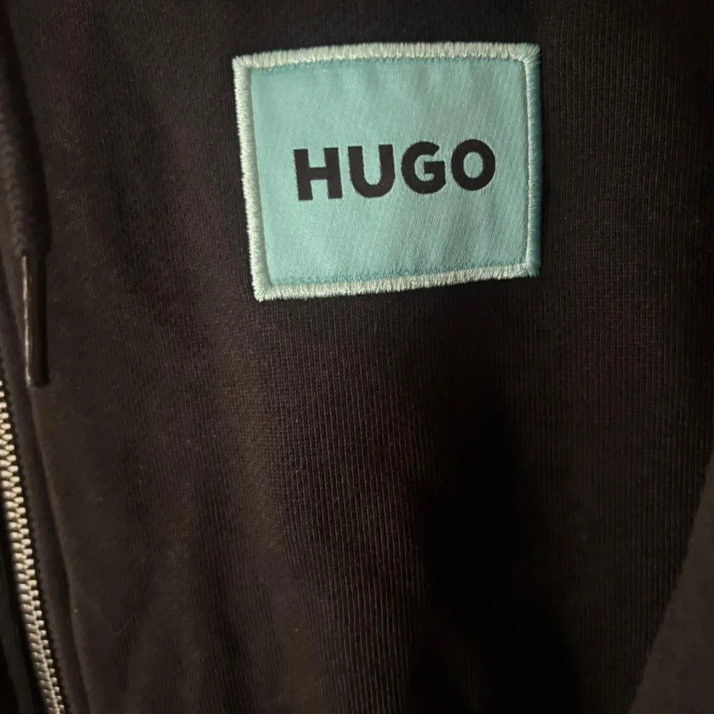 Säljer en svart hoodie från Hugo med dragkedja framtill och en ljusblå patch med logga på bröstet. Tröjan har huva med snörning och ribbade muddar vid ärmslut och nederkant. Perfekt för en avslappnad stil.. Neuletakit & Villapaidat.