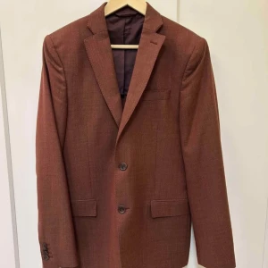 Brun kavaj från J.Lindeberg x Angelico - Säljer en brun kavaj från J.Lindeberg i samarbete med Angelico. Kavajen är slim fit och tillverkad i ull, med två knappar framtill och klassiska fickor. Perfekt för dig som vill ha en stilren och modern look.