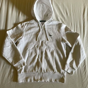 Vit hoodie med dragkedja från Lacoste - Säljer en klassisk vit hoodie från Lacoste med dragkedja och huva. Väldigt ren och fläckfri för att vara en vit hoodie, på sista bilden syns en ytterst liten fläck på höger bakaxel. Nästan osynlig.