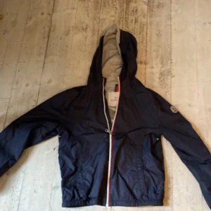 Mörkblå vindjacka från Moncler - Säljer en snygg mörkblå vindjacka från Moncler med huva och dragkedja. Jackan har långa ärmar och en broderad logotyp på ärmen. Perfekt för blåsiga dagar! Andledning att jag säljer är att jag växt ur den. Och den är perfekt nu till våren då den är rätt så tunn.