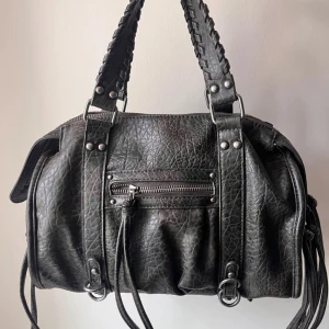 Svart vintage axelväska  - Snygg svart axelväska med coola detaljer. Väskan har en dragkedja framtill och flätade handtag som ger en unik look. Perfekt för att bära med sig det viktigaste med stil. Använd fåtal gånger. 