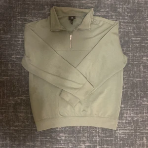 Grön tröja med dragkedja från H&M - Säljer en stilren grön tröja från H&M med dragkedja vid halsen. Tröjan har en relaxed fit och är perfekt för en avslappnad look. Den är långärmad och tillverkad i ett mjukt material.